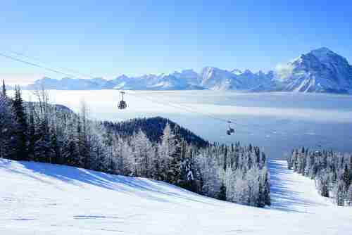 Image is of one of the ski runs at the Lake Louise Ski Resort in Canada. Looking down the ski run into the valley below. Gondolas are also pictured.