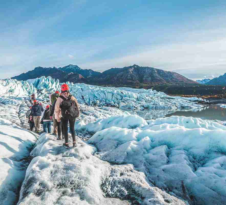 Group hikes across glaciers in Alaska. They have warm clothes and backpacks and face away as they walk in a line across the glacier.